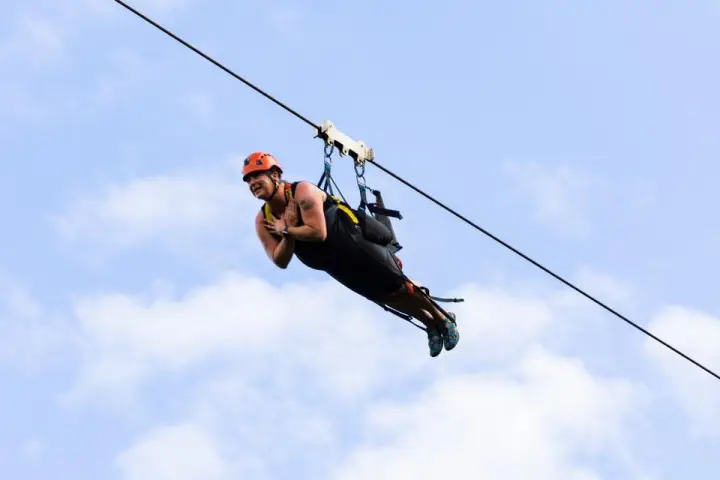 Person ziplining in the sky, wearing a helmet and harness, against a backdrop of clouds.