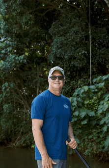 Man in blue shirt and cap holding a paddle by lush green foliage.