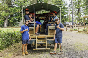 Group of people sitting in a safari vehicle, two standing outside, all wearing helmets in a forest area.
