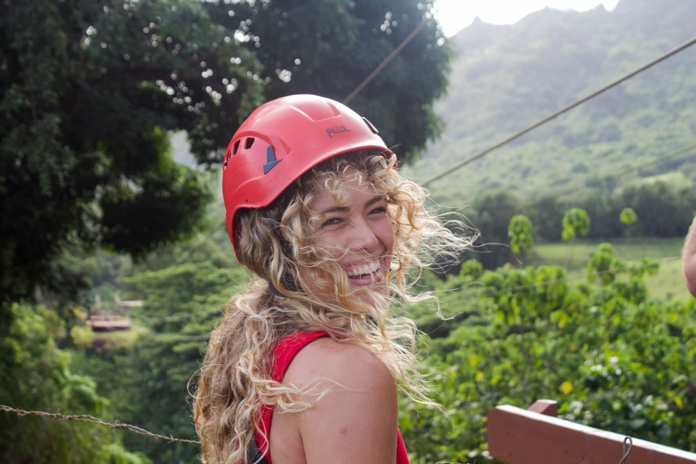 Smiling person with curly hair in a red helmet outdoors ok Kipu Ranch during the Nui Kauai Zipline on Kipu Ranch at Outfitters Kauai.