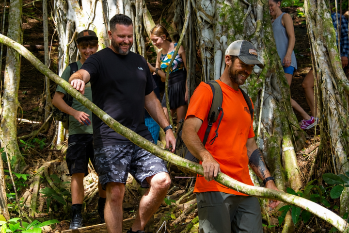 Guests hiking beneath a giant banyan tree on the Kīpū Falls & Treehouse Hike at Kīpū Ranch, Kauaʻi.